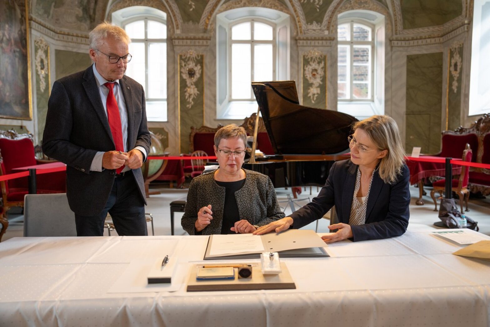 Zwei Frauen sitzen hinter einem Tisch, links neben ihnen steht ein Mann mit Stift in der Hand