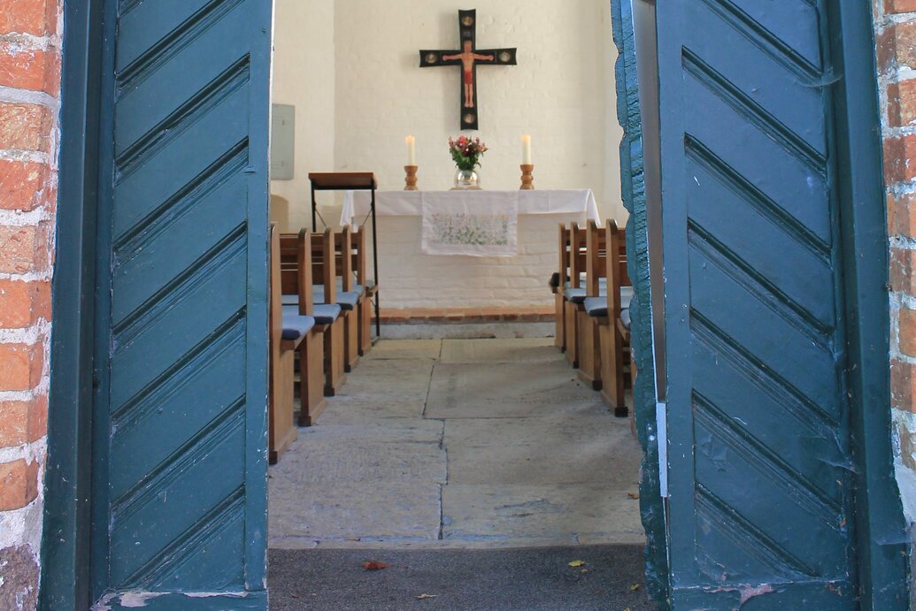 Eine offene blaue Kirchentür gibt den Blick frei zu einem Kirchenaltar auf dem zwei große, weiße, brennende Kerzen zu sehen sind. Zwischen den Kerzen steht eine runde Glasvase mit Blumen, darüber hängt ein Altarkreuz. Im vordergrund sind braune Holz-Kirchenbänke mit blauen Sitzauflagen zu sehen.