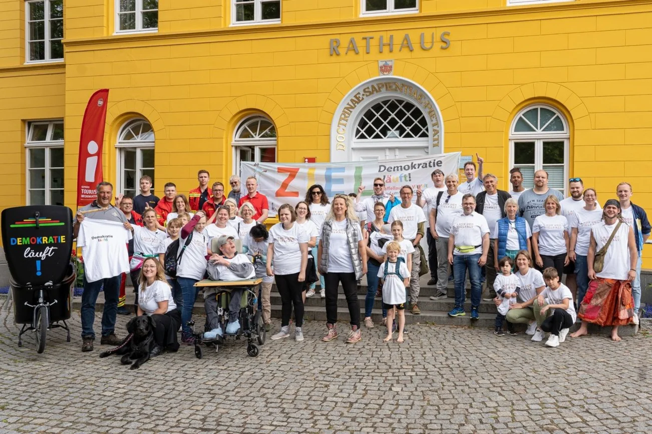 Eine großes Gruppenfoto von Menschen in weißen T-Shirts vor einem gelben Ratzhaus