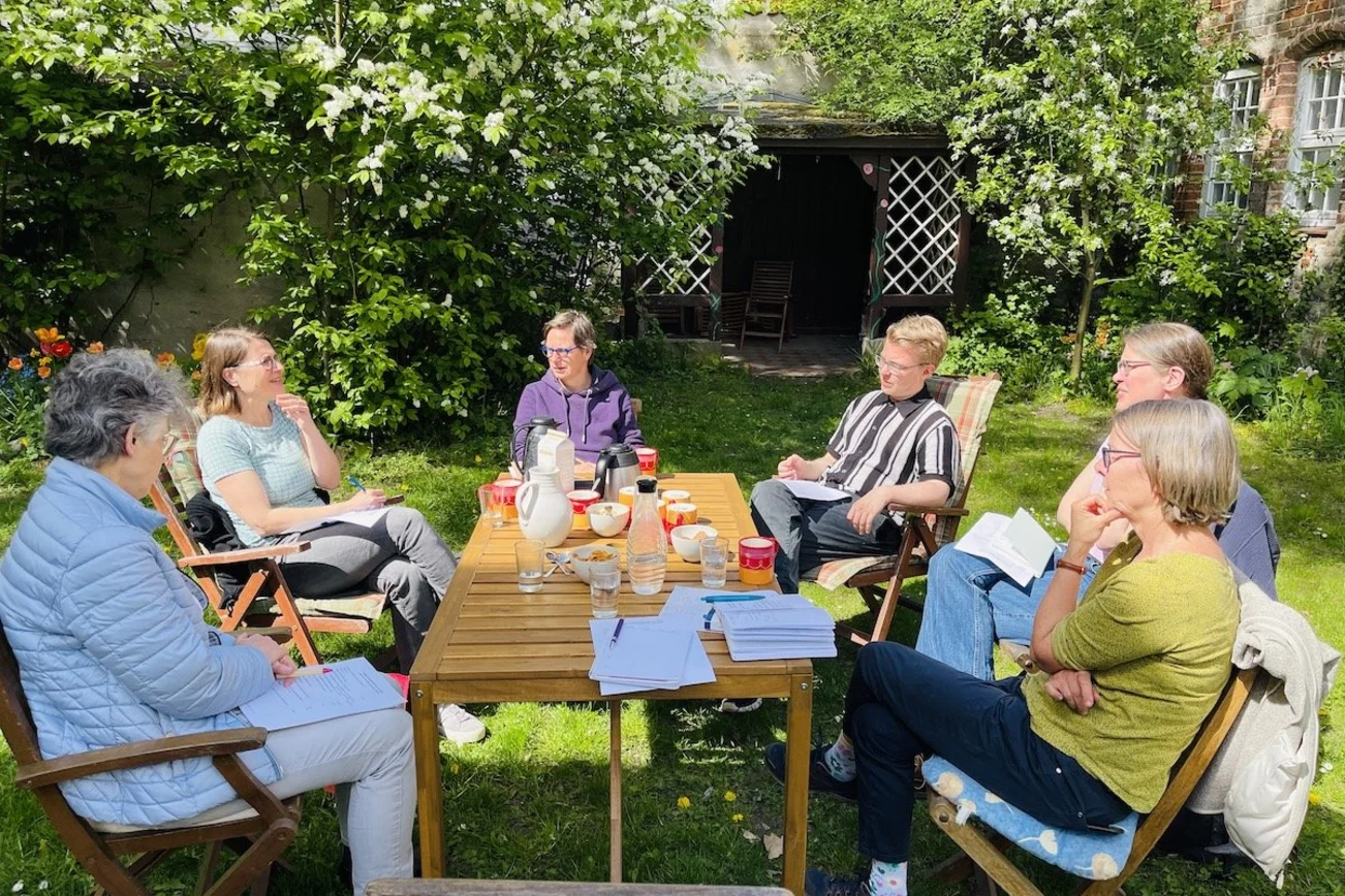 Fünf Pastorinnen und ein Vikar sitzen im sonnigen Garten um einen Tisch und bereiten das Tauffest vor.
