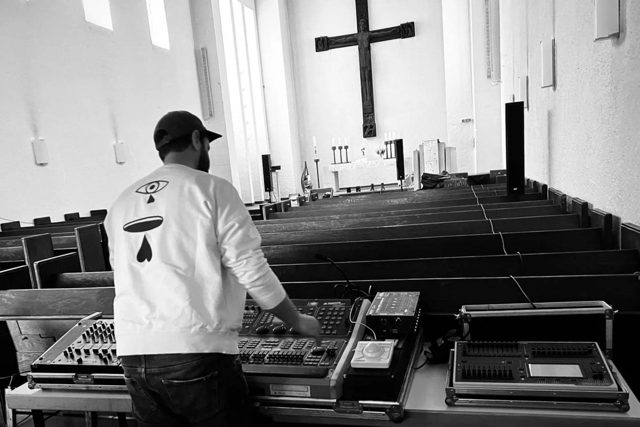 Ein Mann von hinten zu sehen in einem Kirchenraum mit Kreuz und DJ Technik.