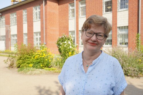 Eine Frau mit kurzen Haaren steht vor einer Hauswand mit Gitterfenstern.