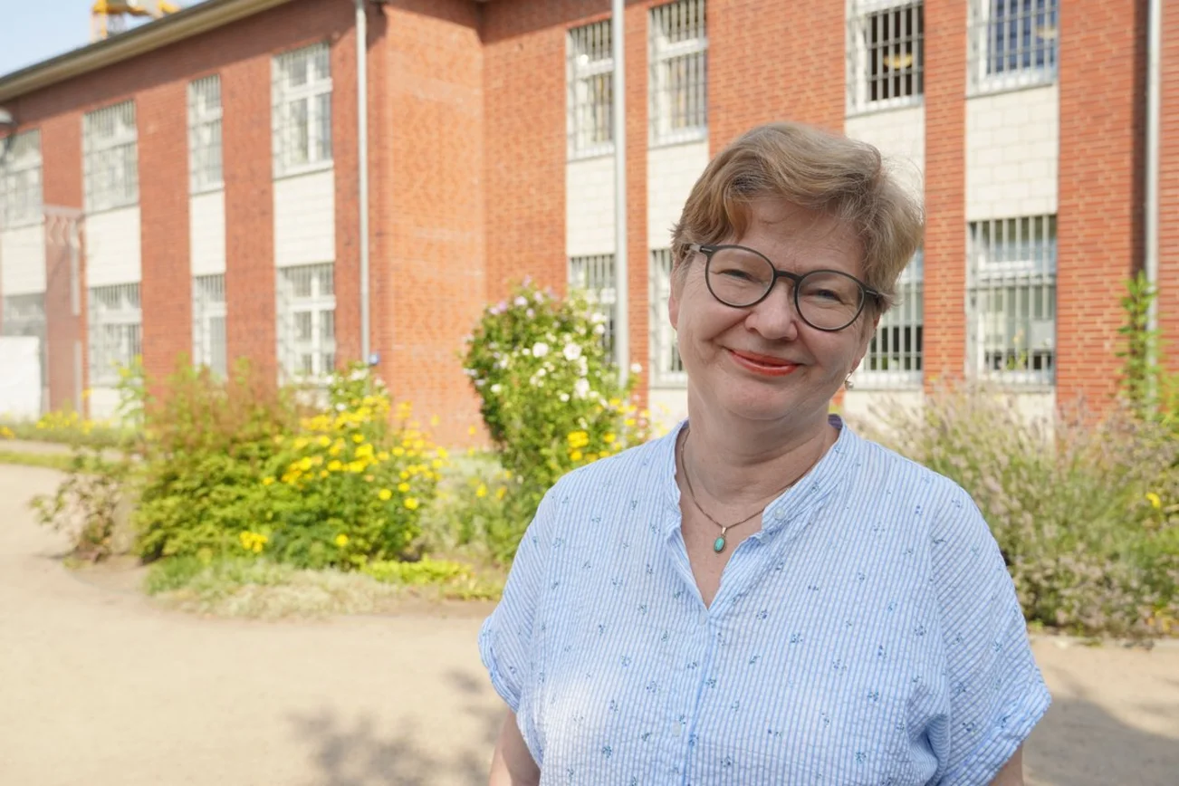 Eine Frau mit kurzen Haaren steht vor einer Hauswand mit Gitterfenstern.