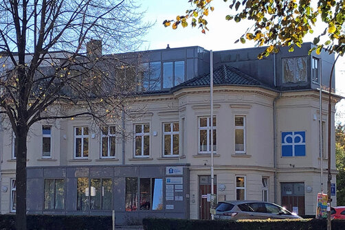 Das schöne beige Altbau-Gebäude der Gemeindediakonie. Im unteren Bereich ist eine Fensterfront zu sehen. Im Vordergrund ein Baum mit wenigen Blättern 