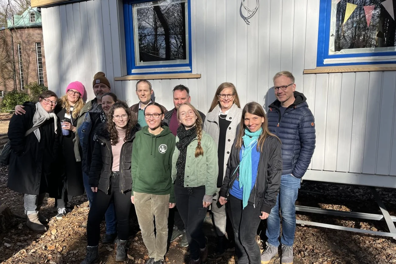 Eine Menschengruppe steht vor einem Bauwagen und lächelt.