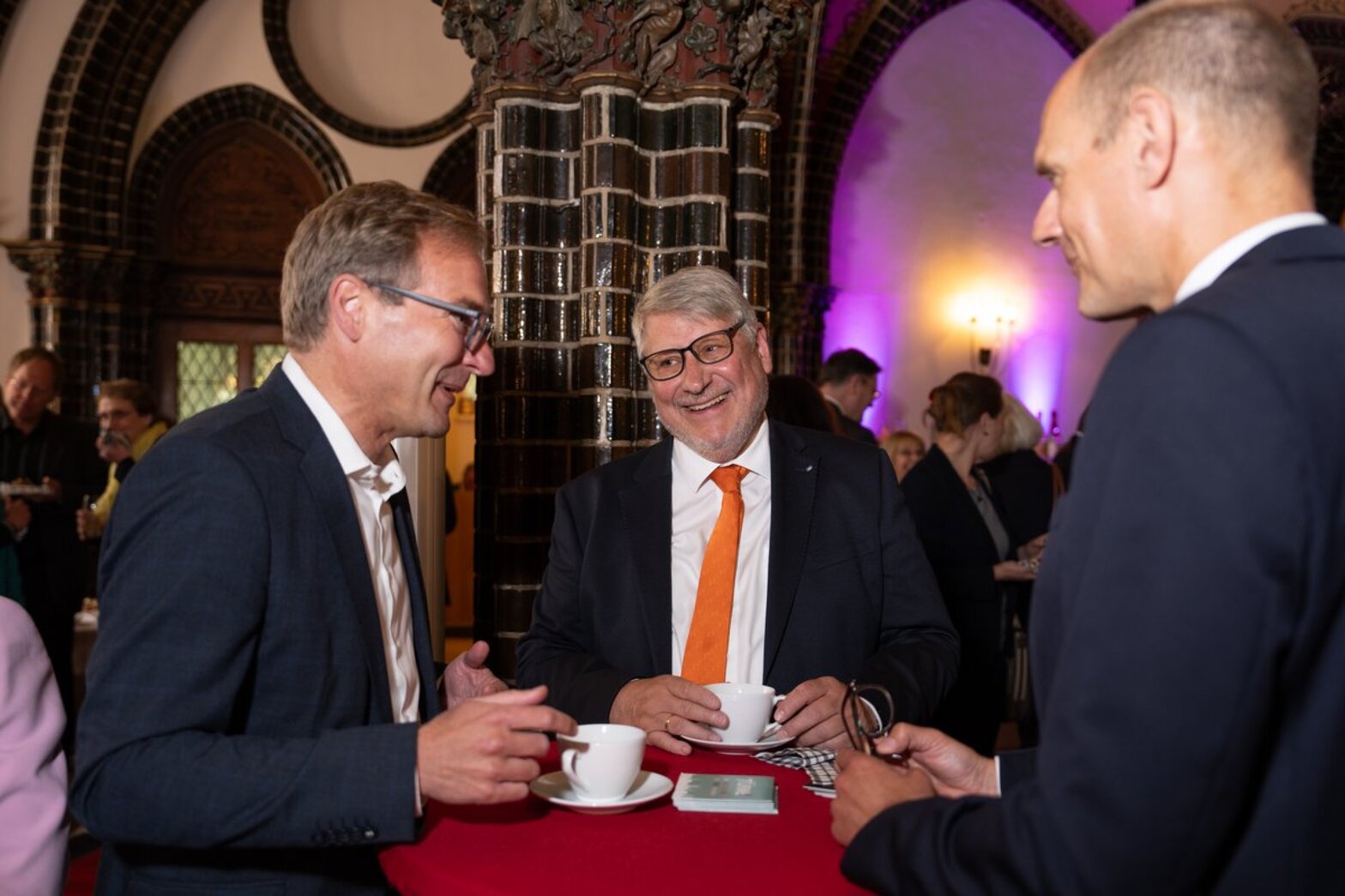 Drei Männer stehen in Anzügen gekleidet vor einer Säule, an einem Stehtisch mit roter Decke und lächeln, zwei weiße Tassen mit Untertassen stehen vor Ihnen.