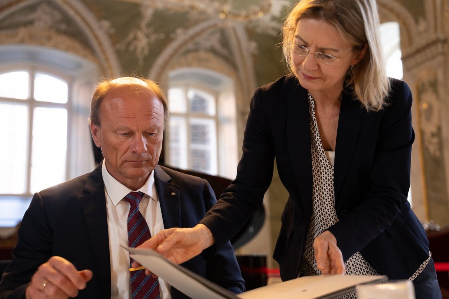 Eine Frau steht und blättert in einer Dokumentenmappe. Links neben ihr sitzt ein Mann und schaut ihr zu.