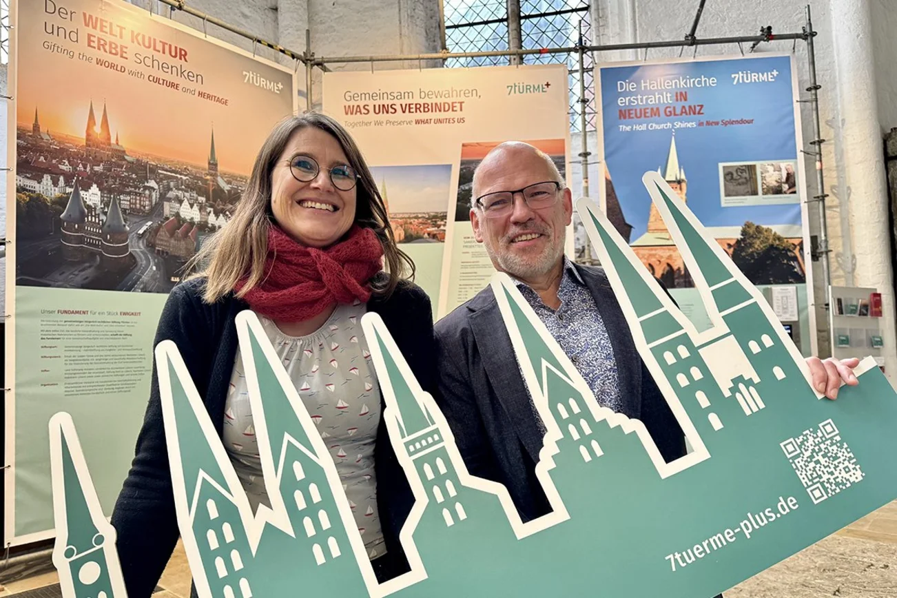 zwei Personen halten einen grünen Austeller mit Kirchtürmen vor sich  - Im Hintergrund sind Banner zu sehen