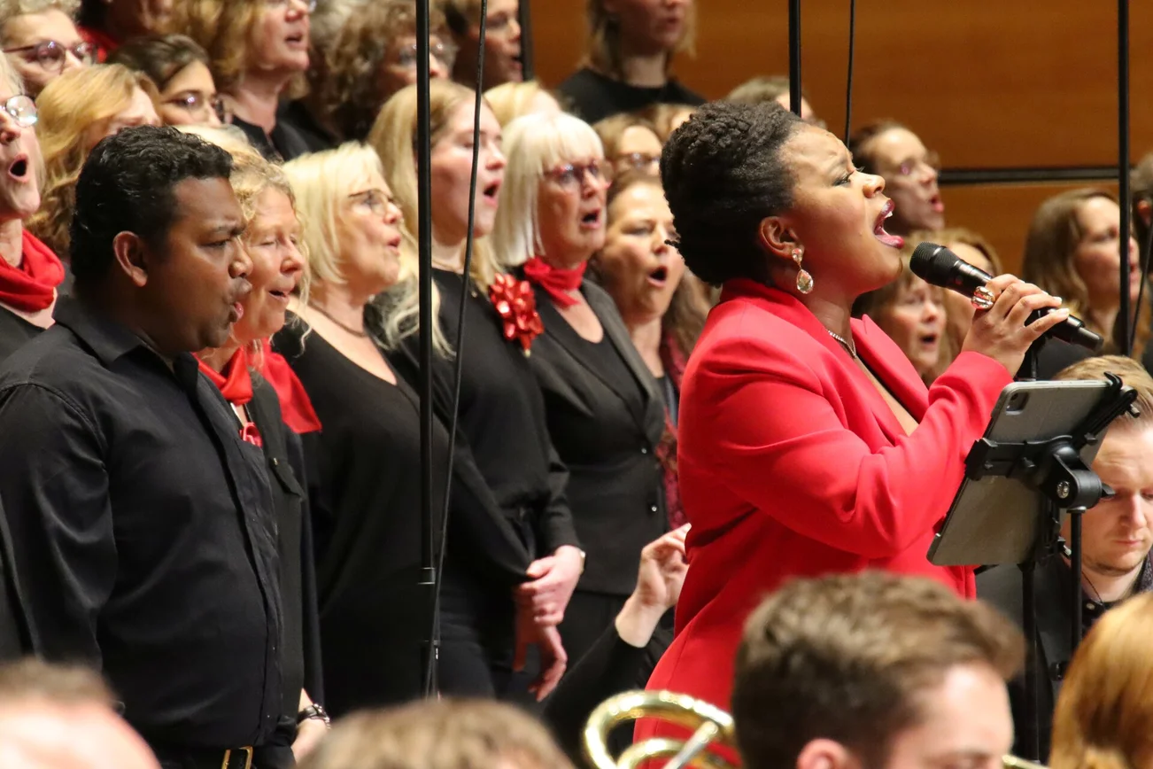 Eine schwarze Sängerin mit rotem Anzug steht inmitten eines Chores und singt. 