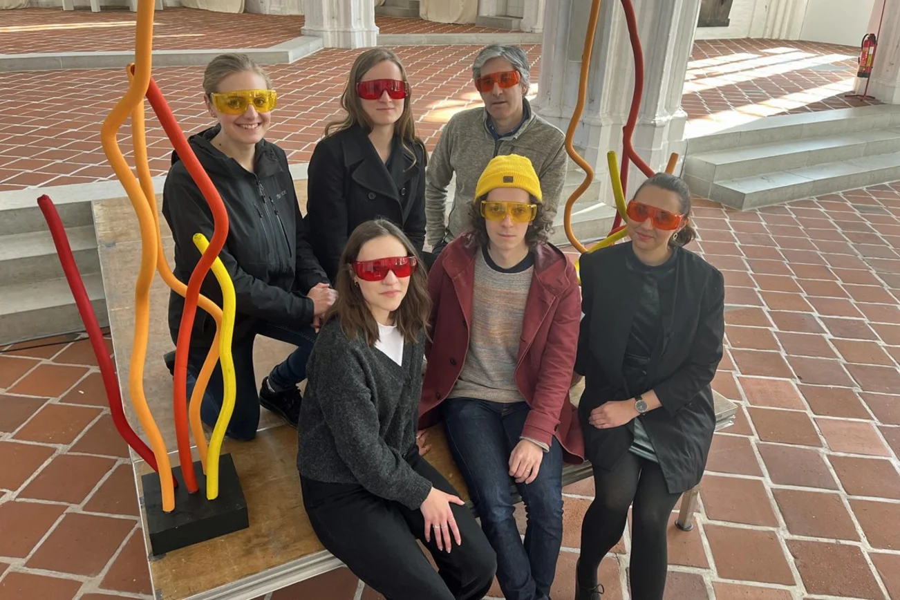 Fünf Personen sitzen auf einem Podest und tragen bunte Schutzbrillen.