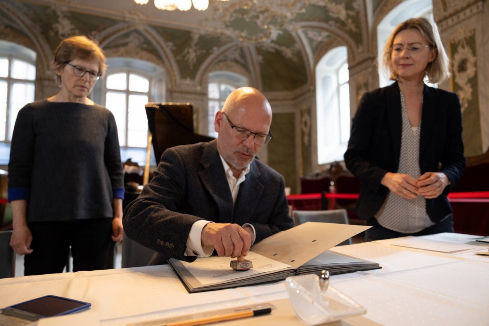 Ein Mann sitzt hinter einem Tisch mit weißer Tischdecke und unterzeichnet ein Dokument, zu beiden Seiten steht eine Frau.