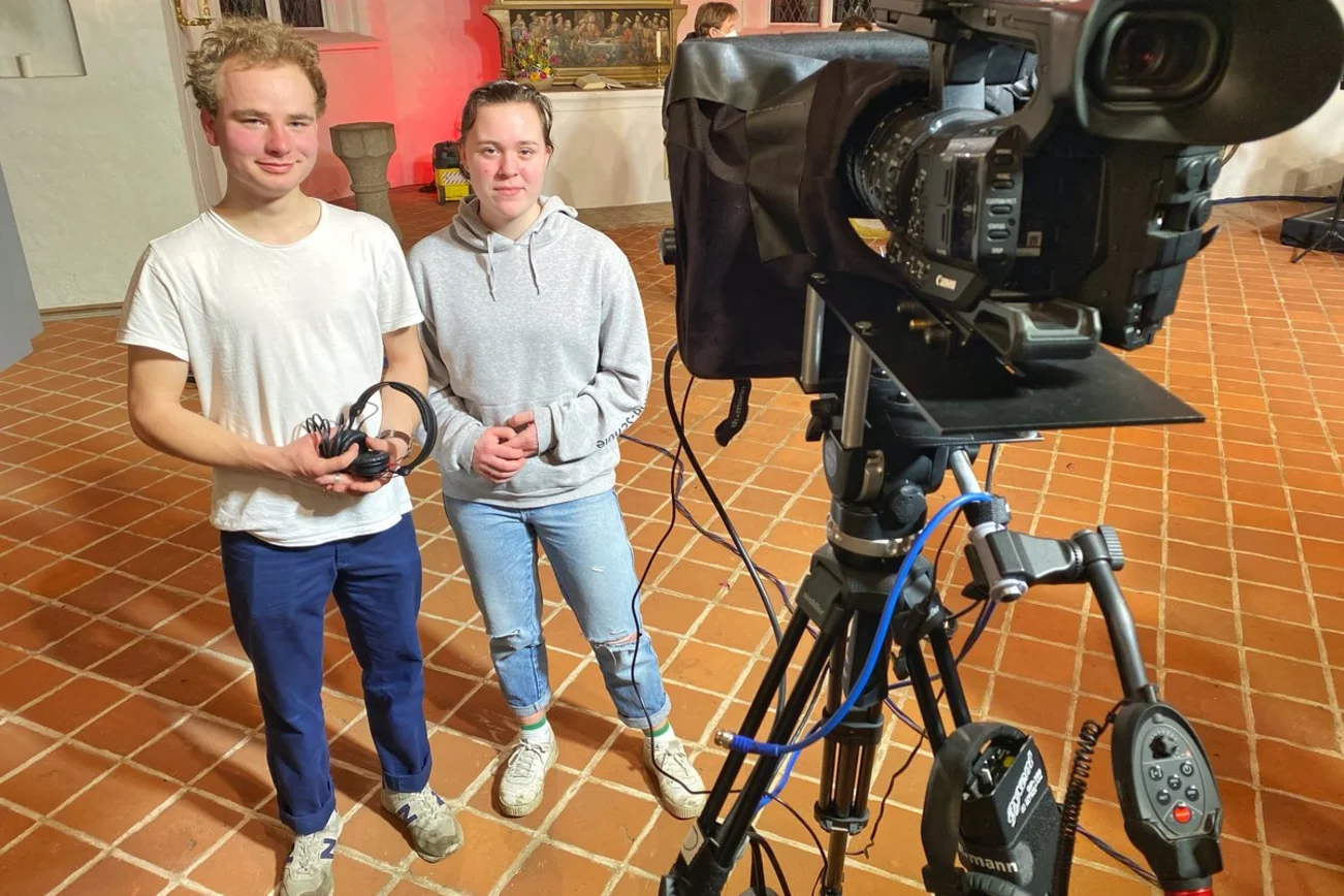 Zwei junge Menschen stehen neben einer Kamera. 