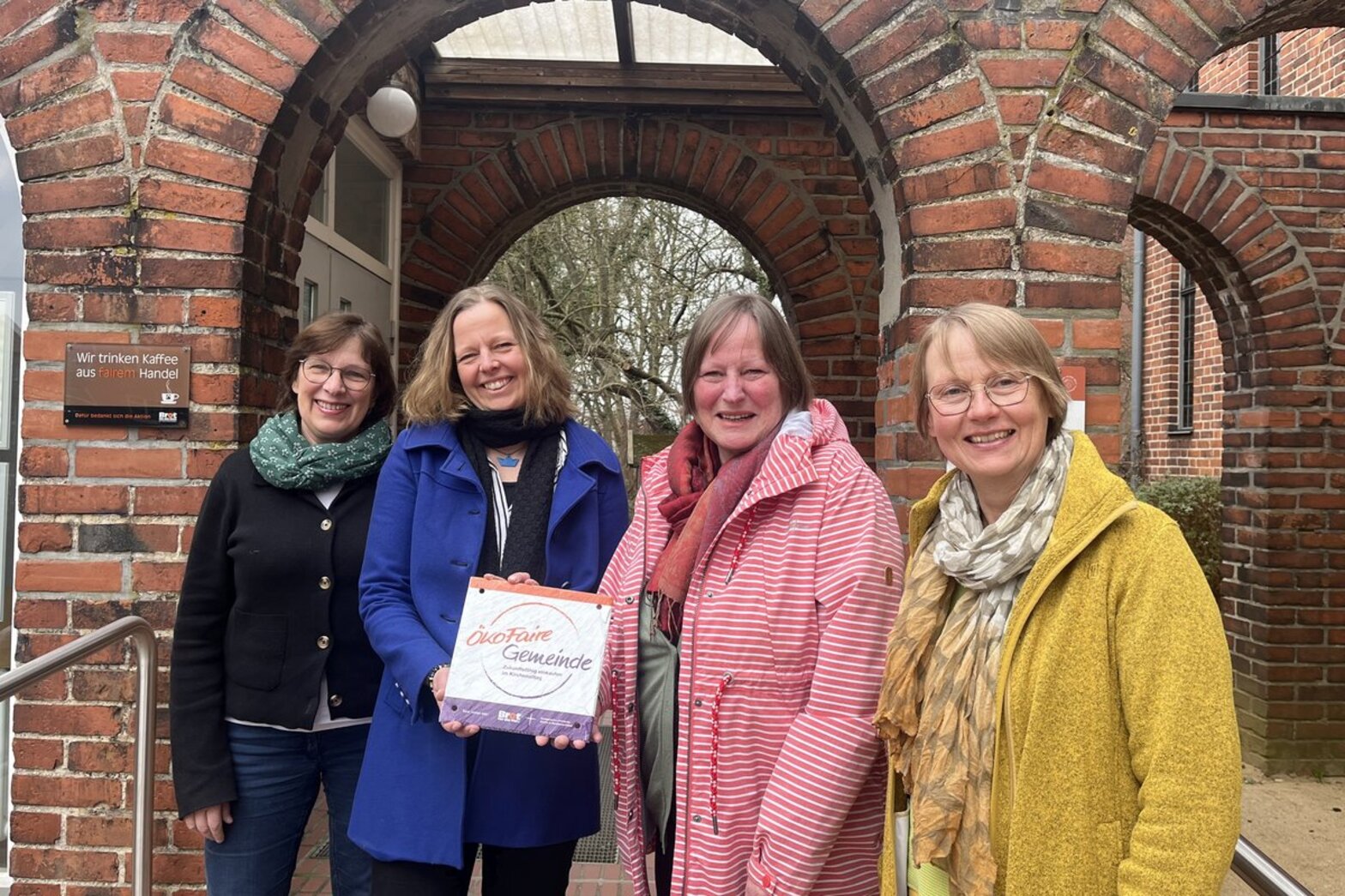 Vier Frauen stehen von Backsteinbögen und halten ein Schild mit der Aufschrift "Ökofaire Gemeinde"