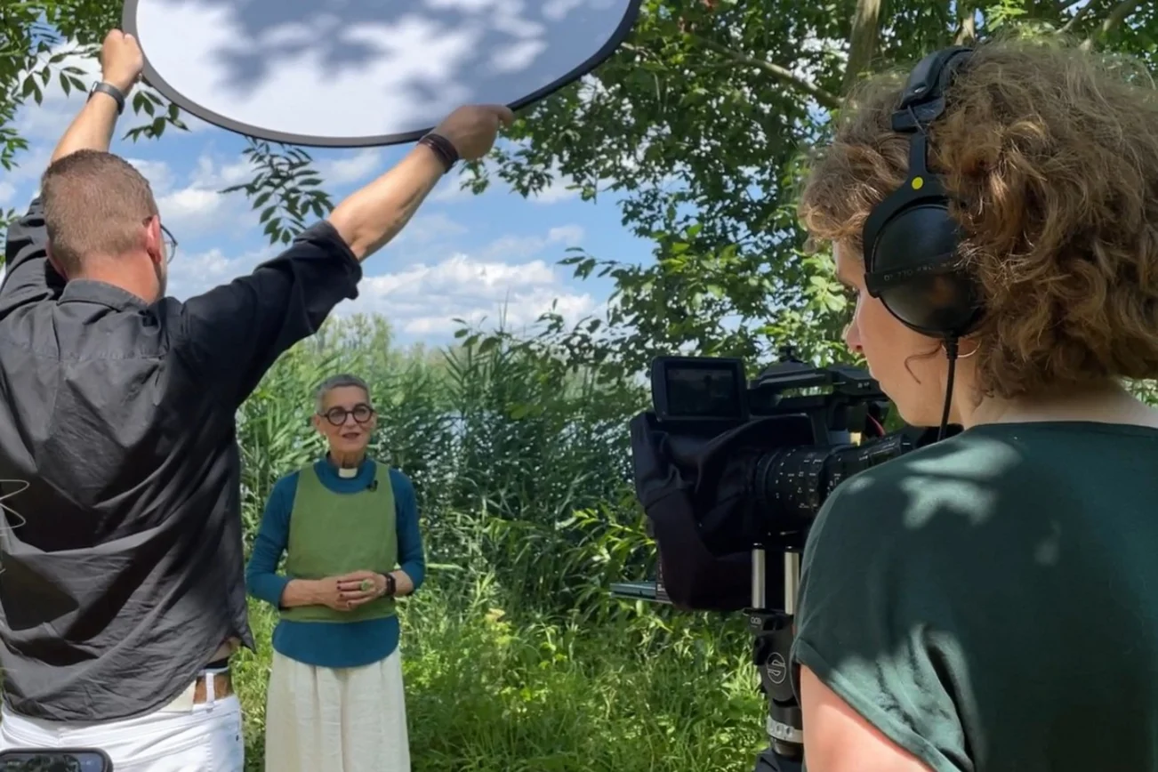 Pastorin Katja von Kiedrowski wird in einer üppigen Naturlandschaft gefilmt. Eine Kamera zeigt in ihre Richtung.