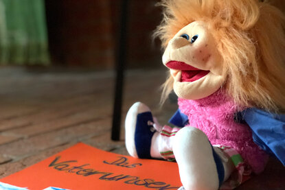 Eine Handpuppe mit langen Haaren, sitzt rechts in der Ecke auf einem roten Pappschild, auf welchem 'Das Vaterunser' steht - Copyright: Ev.-Luth. Kirchenkreis Lübeck-Lauenburg