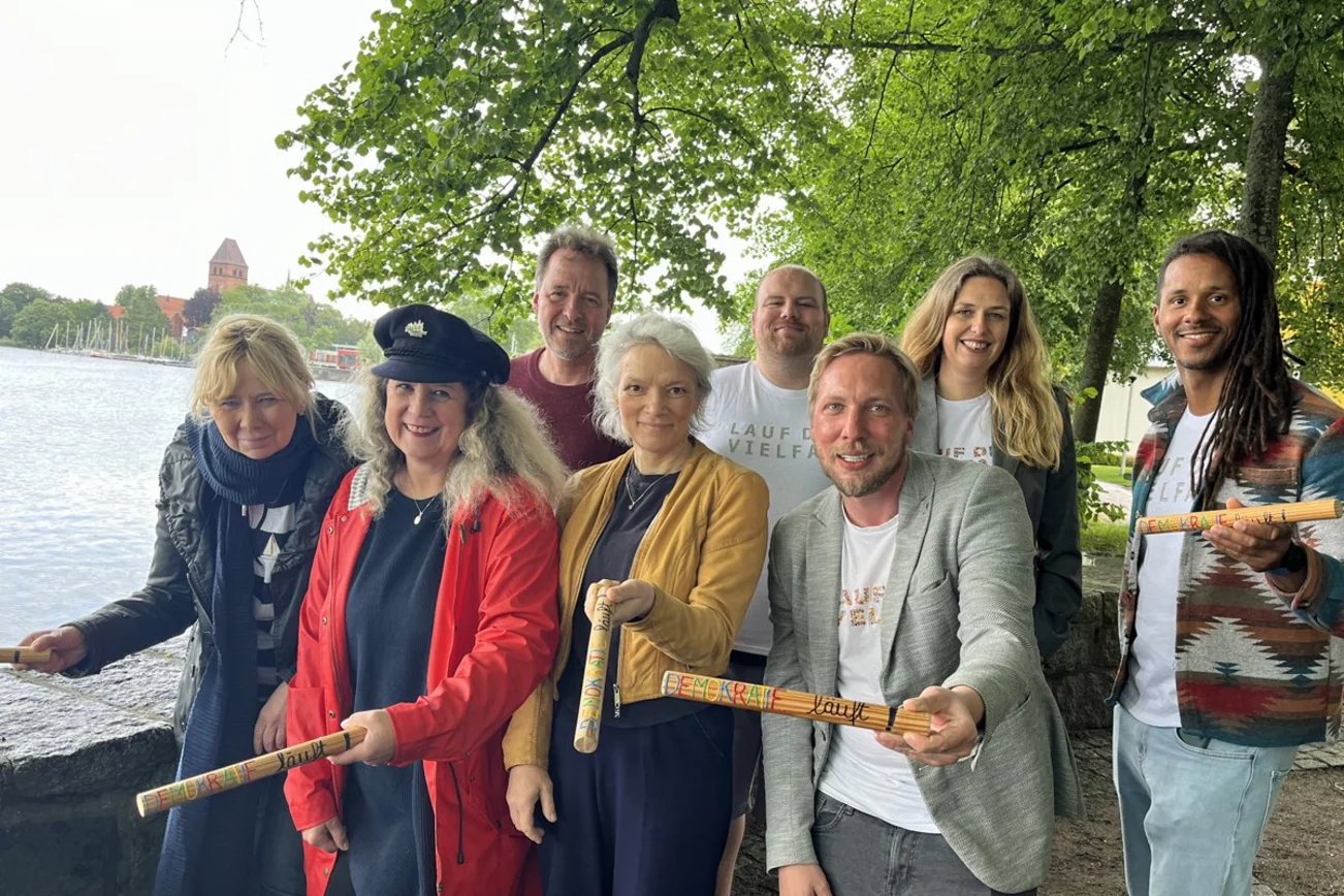 Mehrere Menschen stehen an einem See und halten Staffelstäbe in den Händen.