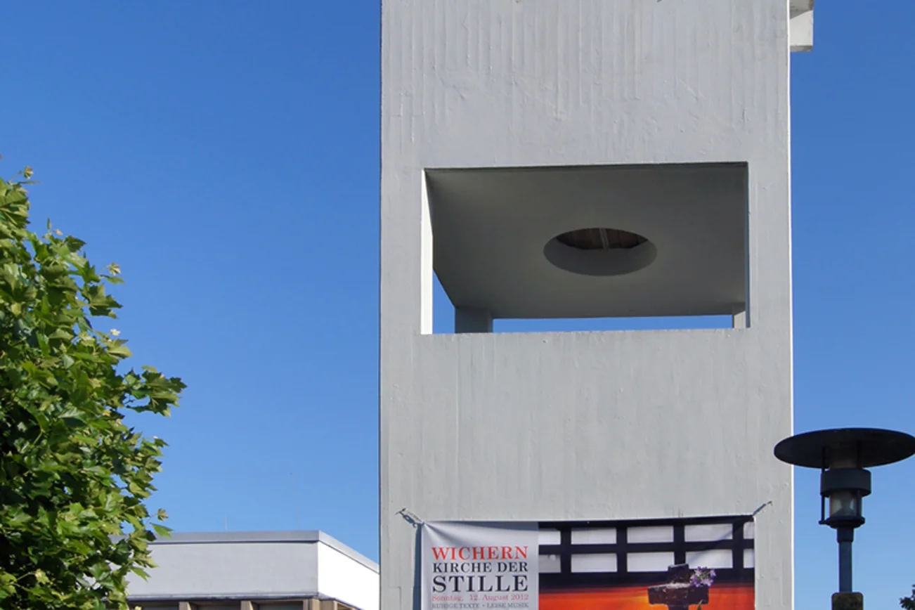 Beton-Glockenturm mit Spannband 'Kirche der Stille', darunter Schatten, links Ecke des Kirchsaals mit Eingang, am Rand grüne junge Linde, rechts eine Laterne