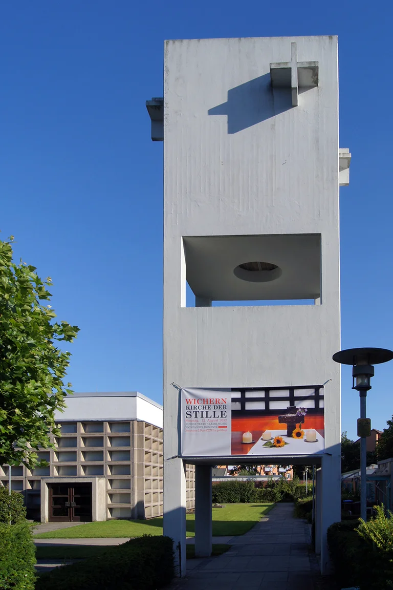 Beton-Glockenturm mit Spannband 'Kirche der Stille', darunter Schatten, links Ecke des Kirchsaals mit Eingang, am Rand grüne junge Linde, rechts eine Laterne