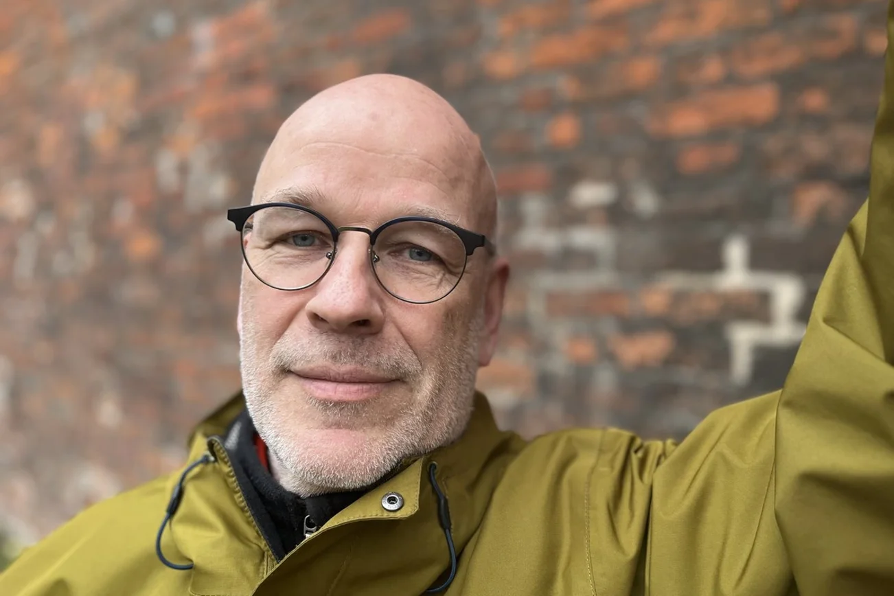 Ein Mann steht vor einer Backsteinmauer und schaut in die Kamera.