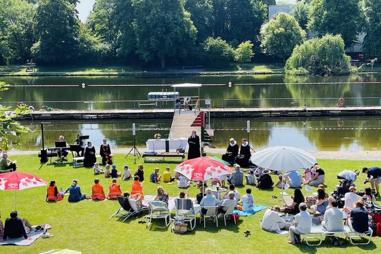 Blick von oben in das Altstadtbad Krähenteich im Jahr 2023. Es sind viele Familien zu sehen, die im Sonnenschein sitzen und den Gottesdienst mit den Pastor:innen mitmachen.
