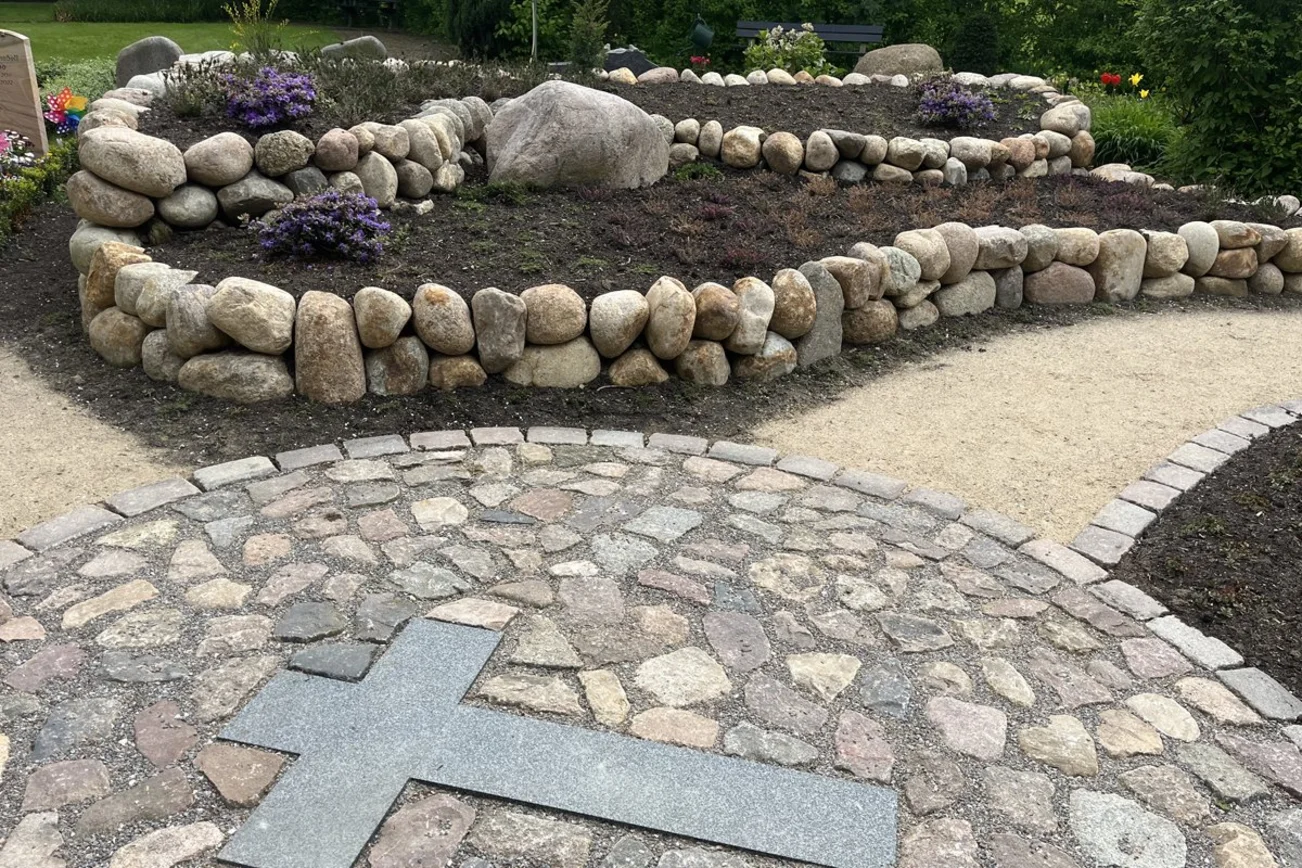 Ein in den Boden eingelassenes Kreuz vor einer mit Steinen umfassten Grabanlage