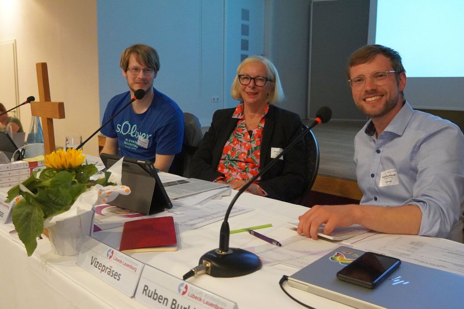 Eine Frau und zwei Männer sitzen an einem Tisch mit Mikrofonen und einem Holzkreuz.