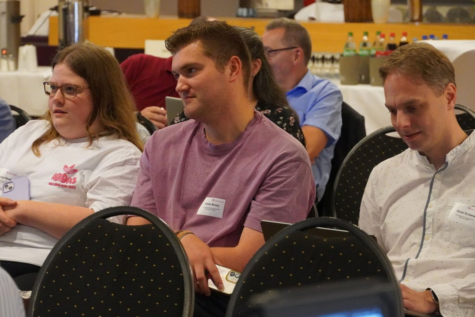 Drei Personen sitzen in einer Stuhlreihe und hören zu. 