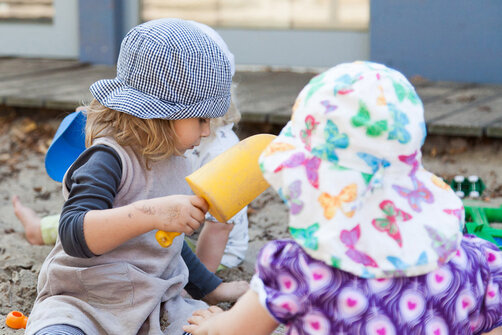 Zwei kleine Kindergartenkinder sind in einem Sandkasten und spielen. Beide Mädchen tragen Sonnenhüte. Das Mädchen links hält eine Schaufel in der Hand.