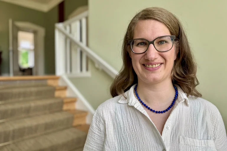 Eine Frau mit langen blonden Haaren und einer dunklen Brille lächelt in die Kamera. Hinter ihr ist eine Treppe zu sehen, so wie hellgrüne Wände.