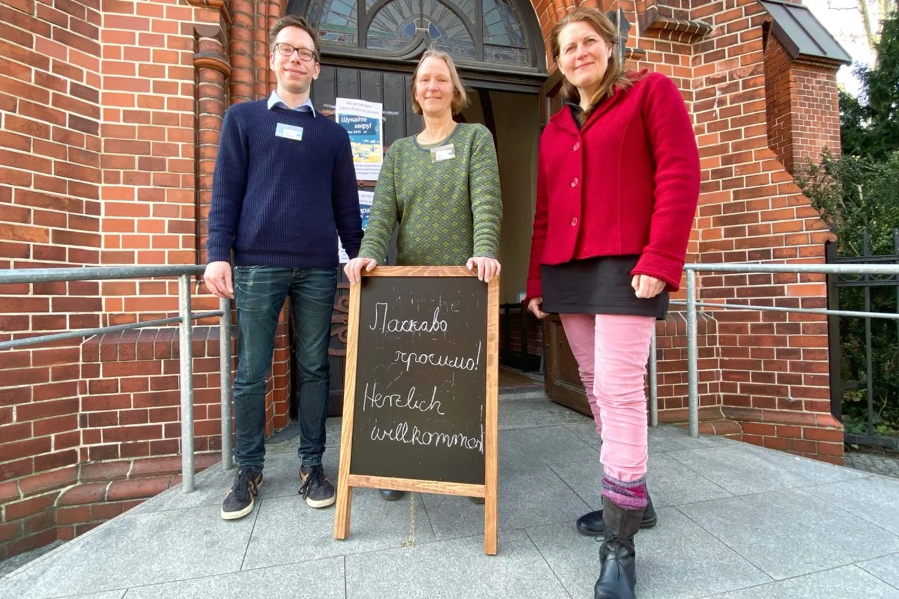 Drei Personen stehen vor dem Eingang einer Kirche. 