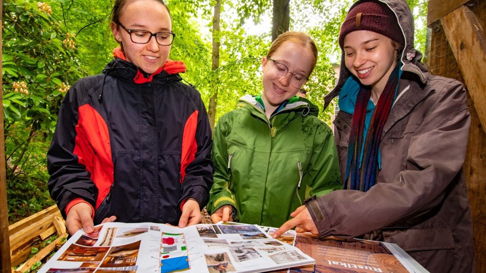 Drei junge Frauen schauen sich Pläne und Fotos an. Sie stehen im freien, unter grünen Bäumen. 