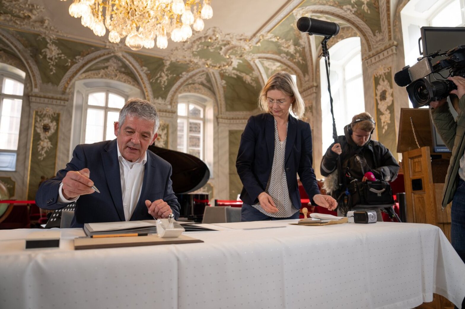 Ein Mann sitzt hinter einem Tisch und will ein Dokument unterschreiben. Eine Frau steht neben ihm und neben ihr ein Tontechniker, welcher ein Mikrofon an einem Stab hält | rechts an der Seite sieht man ein Teil von einer Kamera