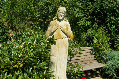 Stein-Statur neben einer Holzbank zwischen gründen Pflanzen und Bäumen - Copyright: Ev.-Luth. Kirchenkreis Lübeck-Lauenburg
