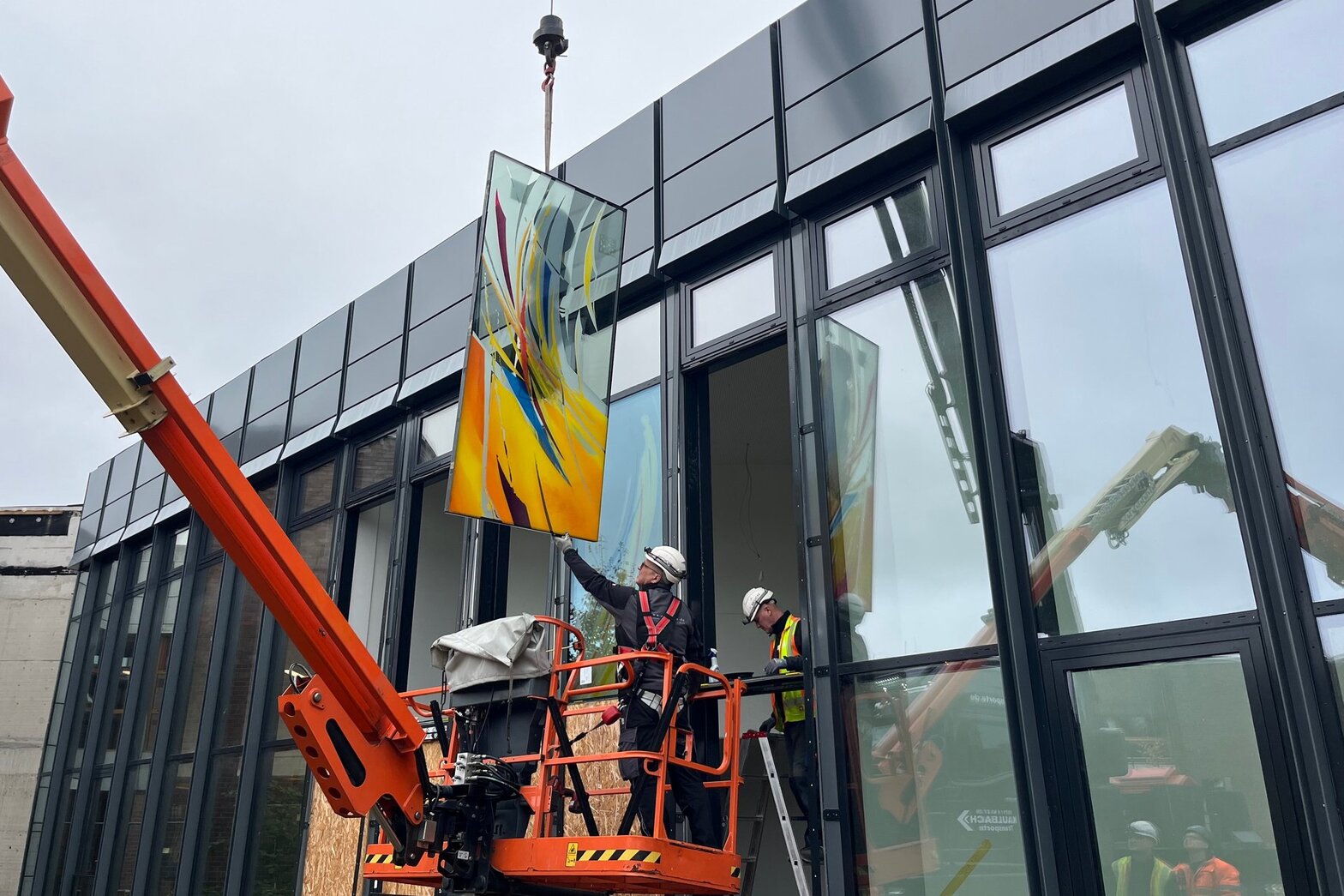 Ein Monteur in Sicherheistkleidung nimmt ein großes buntes Fenster entgegen.