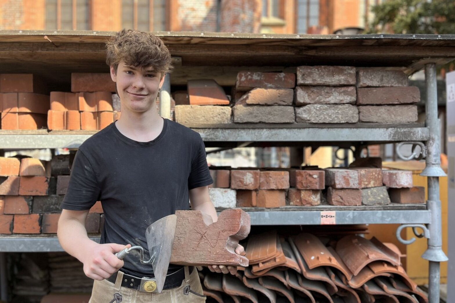 Ein junger Mann steht vor einem Regal mit Ziegeln und hält eine Maurerkelle und einen Ziegelstein in den Händen.