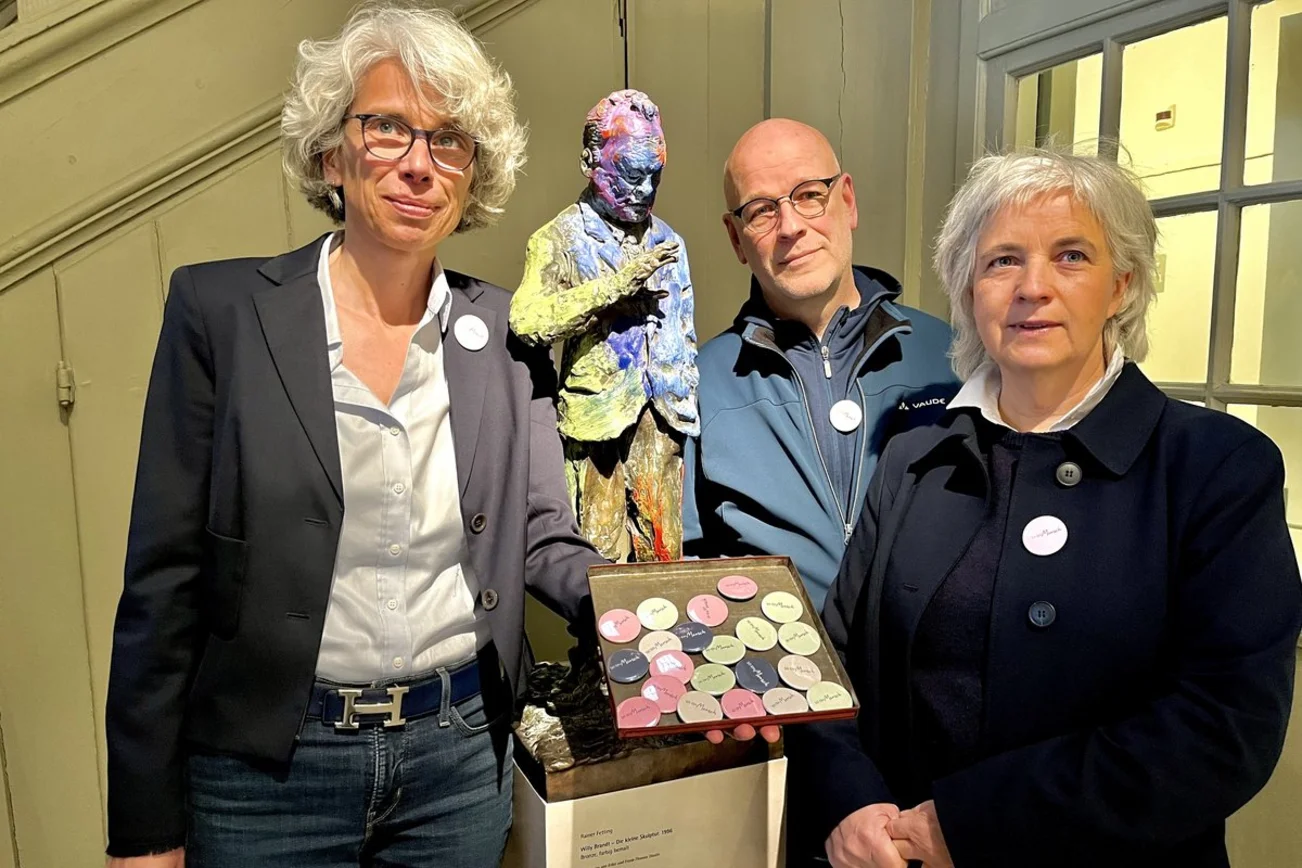 Zwei Frauen und ein Mann stehen neben einer Skulptur von Willy Brandt.