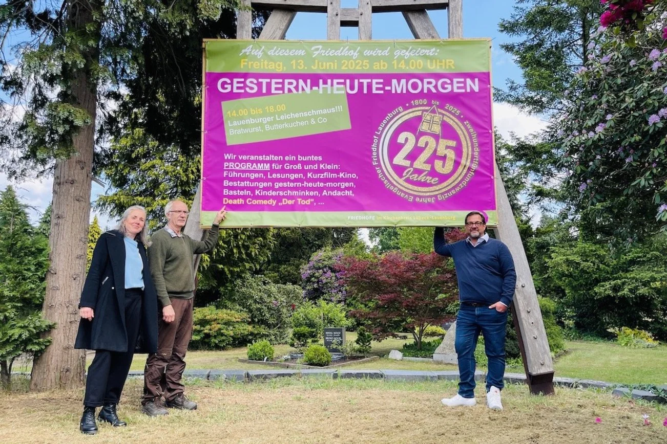 Auf dem Bild stehen Verwaltungsleitung Annett Topaloğlu, der gärtnerische Leiter des Friedhofs, Uwe Pusback, und der Friedhofsbeauftragter im Kirchenkreis Lübeck-Lauenburg, Bernd C. Jakob, vor einem überdimensionalen großen Schild in lila mit der Einladung zum 225. Geburtstags des Lauenburger Friedhofs. 
