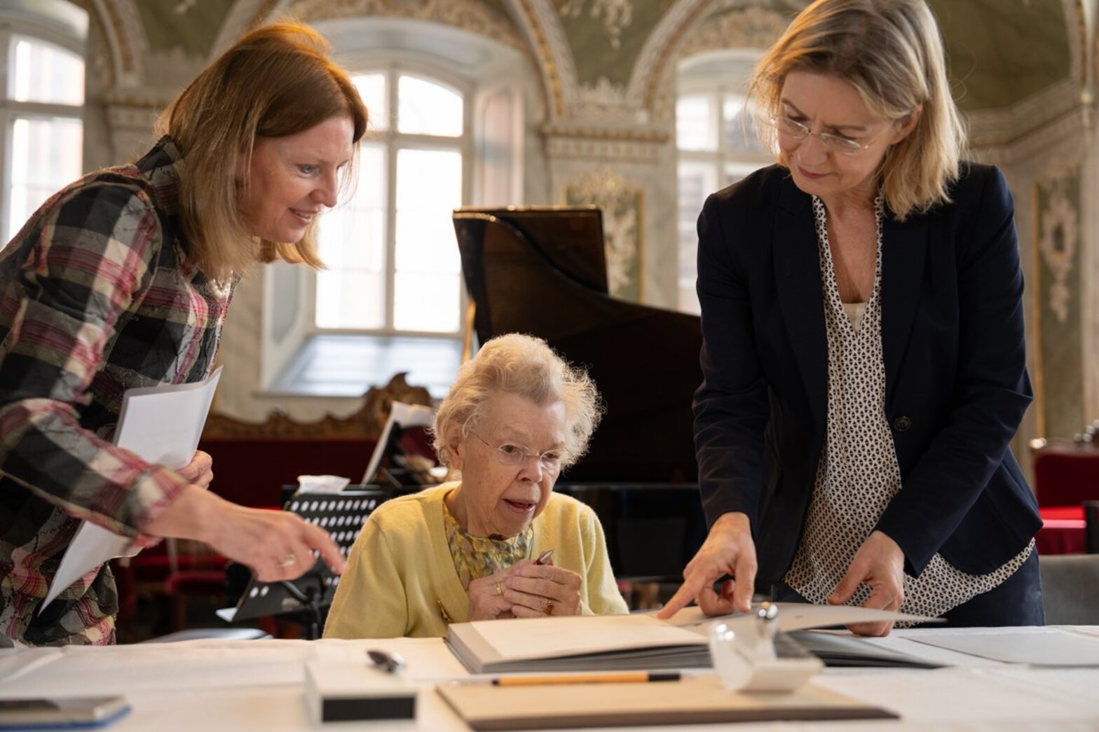 Eine ätere Dame sitzt hinter einem Tisch, rechts von ihr zeigt eine stehende Frau auf ein vor ihr liegendes Dokument, eine weitere Frau steht links hinter dem Tisch