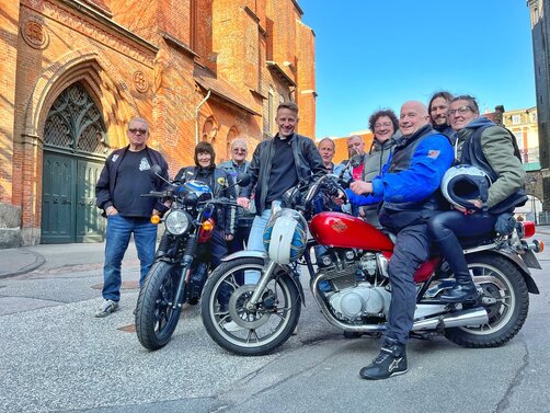 Eine Gruppe Menschen stehen mit zwei Motorrädern vor einer Kirche. 