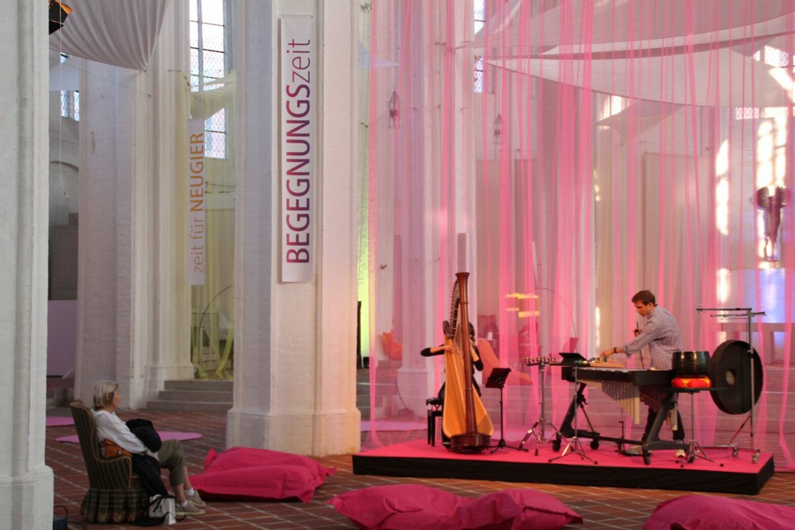 Zwei Personen musizieren auf Harfe und Marimba in einer mit Tüchern behängten Kirche. 
