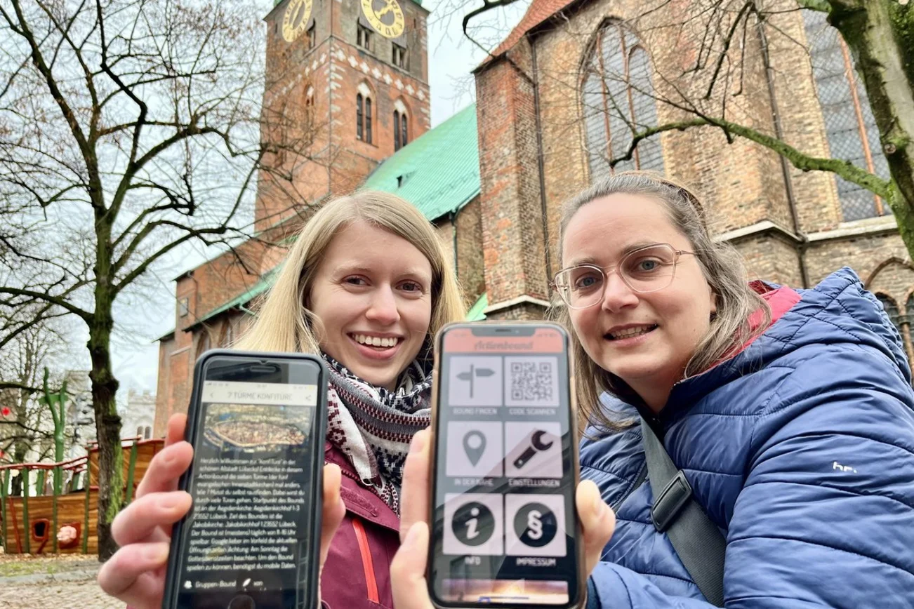 Zwei Frauen stehen vor einer Kirche und halten Smartphones in die Kamera. 