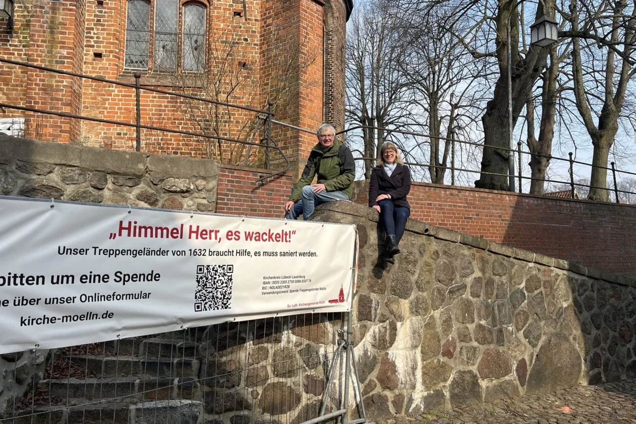 Alte Kirchenmauer mit einem Spendenbanner und zwei Personen, die auf der Mauer sitzen.