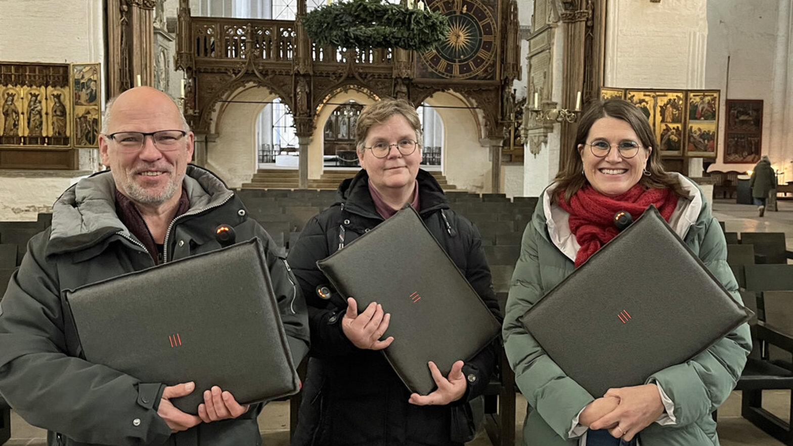 Ein Mann und zwei Frauen stehen in einer Kirch und halten graue Sitzkissen in den Händen