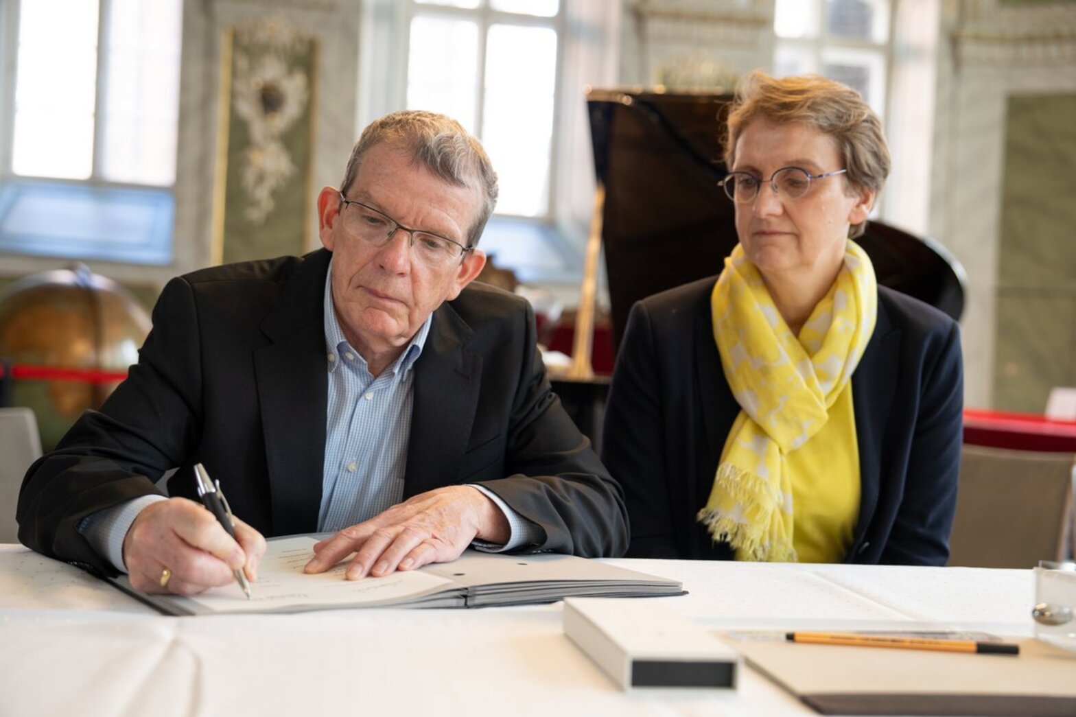 Ein Mann sitzt hinter einem Tisch und unterzeichnet ein Dokument, eine Frau, rechts neben ihm, mit gelbem Schal, schaut ihm dabei zu.