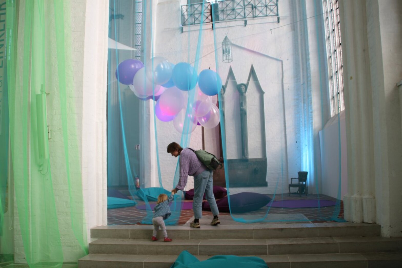 Eine erwachsene Person steht in einer mit Tüchern und Luft behängten Kirche und hält ein Kleinkind an der Hand.