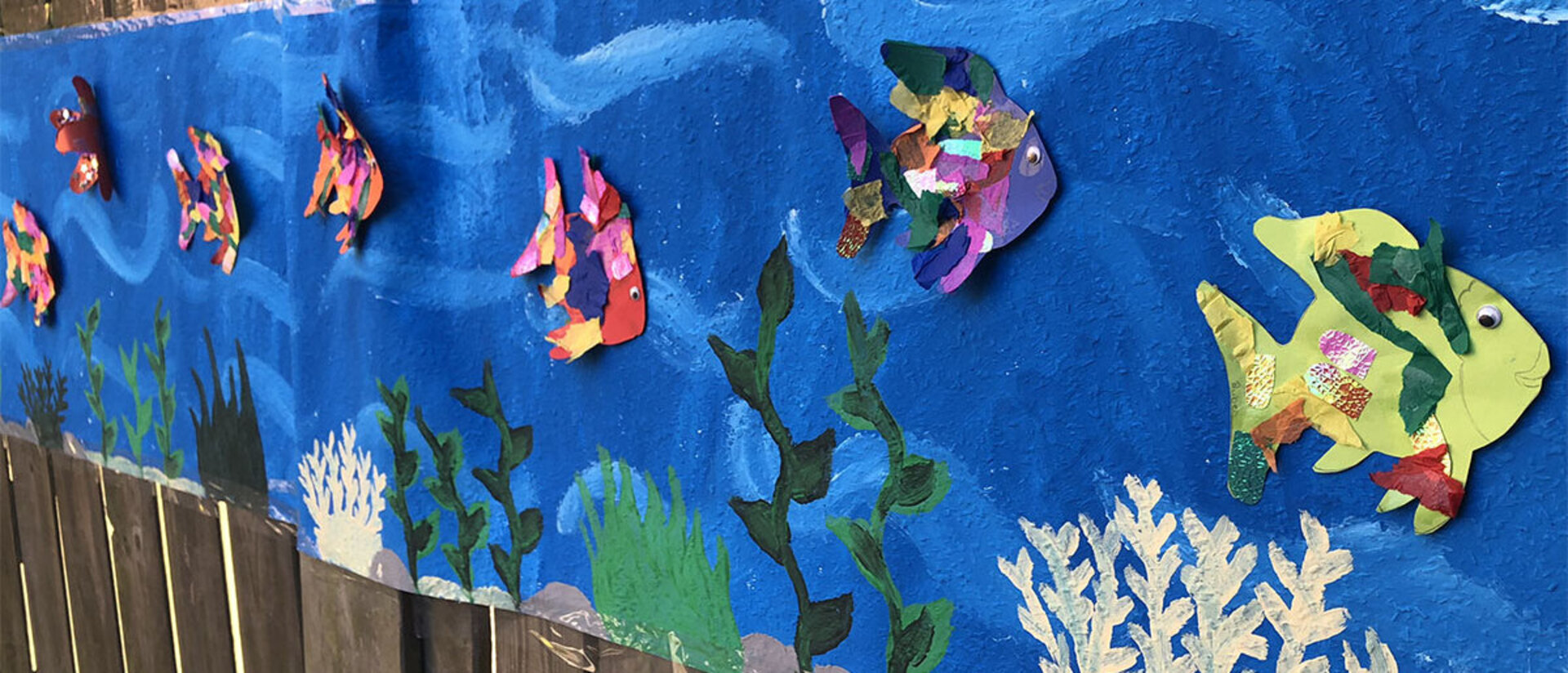 Aus Papier gebastelte bunte Fische auf einem langen blauen Papierstreifen angebracht, welches mit Wasserpflanzen bemalt ist. Der Streifen ist an einem braunen Lattenzaun befestigt.