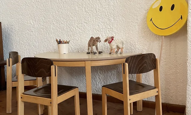 Kleine Kinderstühle um einen kleinen Kindertisch - Gegen die Wand gelehnt ein gelber Holz-Smily