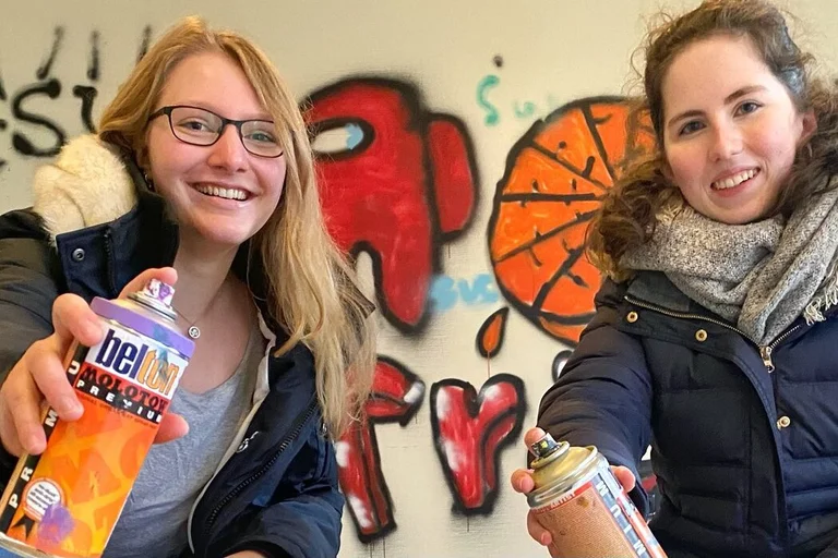 Zwei junge Frauen lächeln mit einer Farbsprühflasche in der Hand in die Kamera.