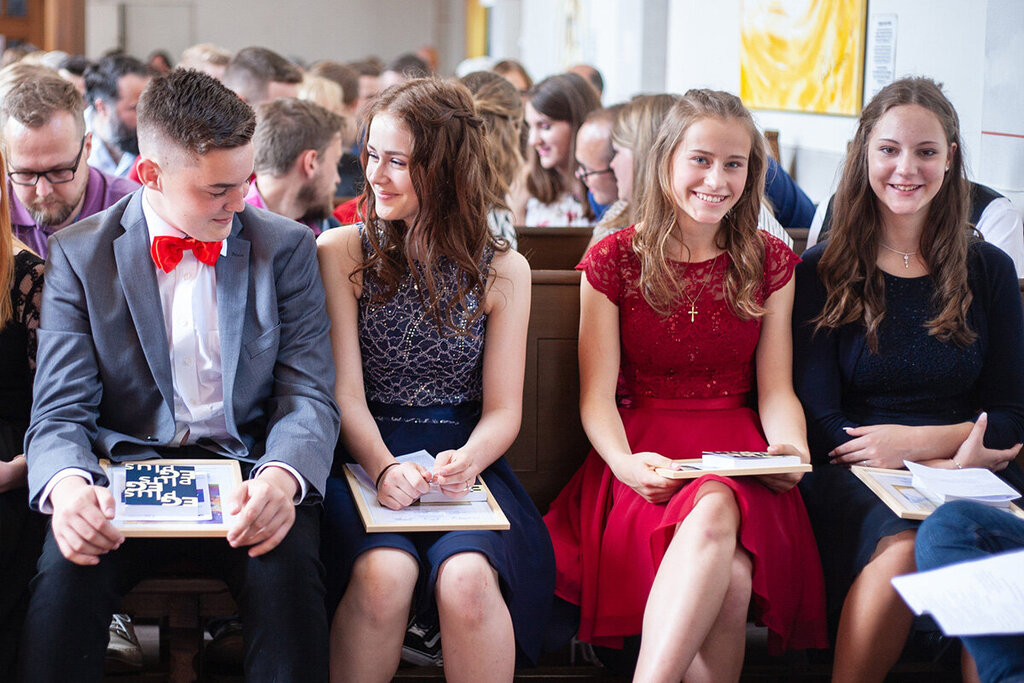 Ein Teenagerjunge mit blau-grauem Sakko, weißem Hemd und roter Fliege sitzt neben drei Teenager-Mädchen. Die Mädchen haben alle lange Haare und sie tragen festliche Kleider. Sie sitzen auf Holzbänken in einer Kirche. Hinter Ihnen sitzen noch mehr Leute auf Holzbänken in mehreren Reihen