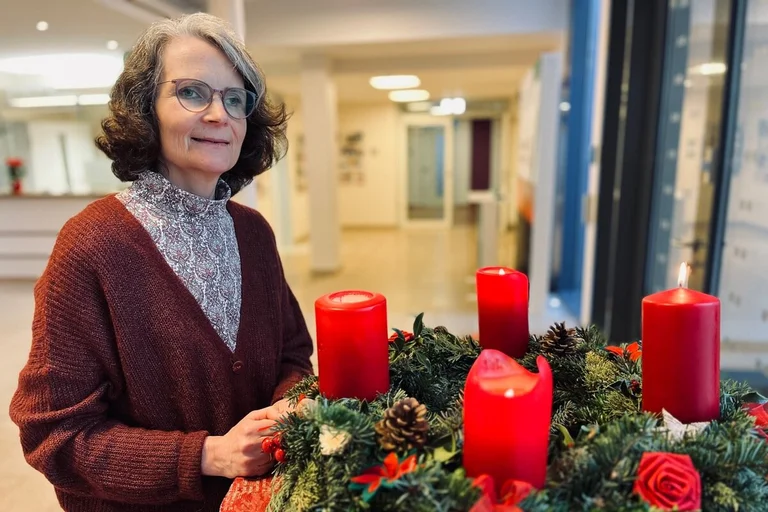 Frau steht lächelnd neben einem Adventskranz an dem vier Kerzen angezündet sind.